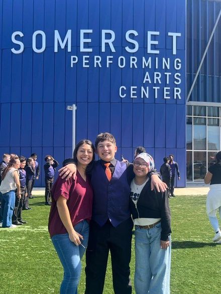 Three people smile and pose in front of the blue Somerset Performing Arts Center building on a sunny day.