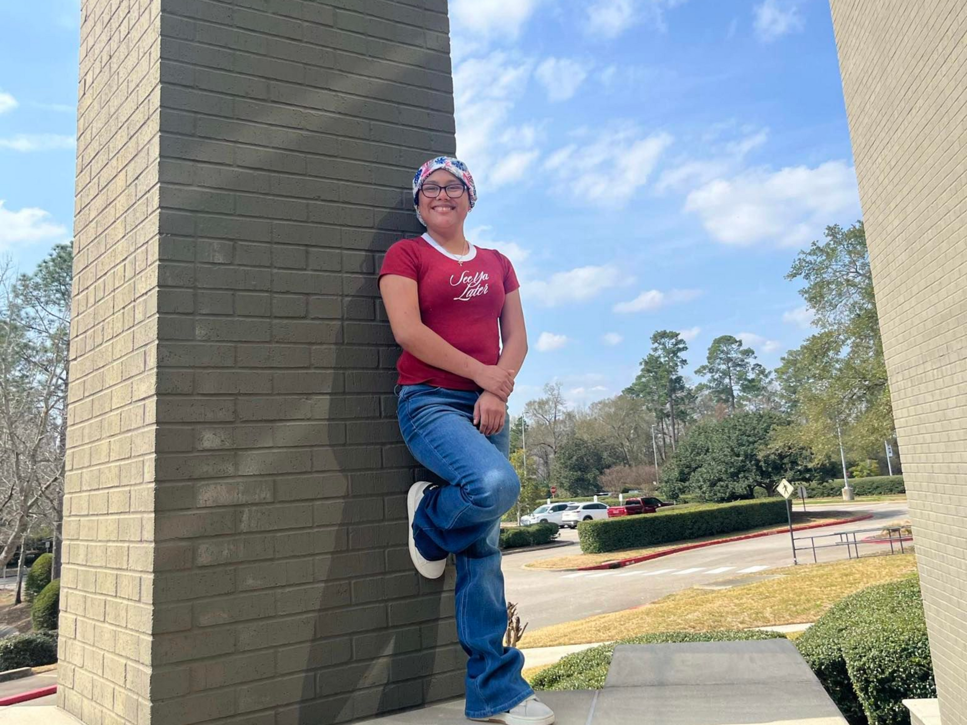 A smiling person wearing a red shirt, blue jeans, and a patterned headscarf leans against a brick pillar outdoors.