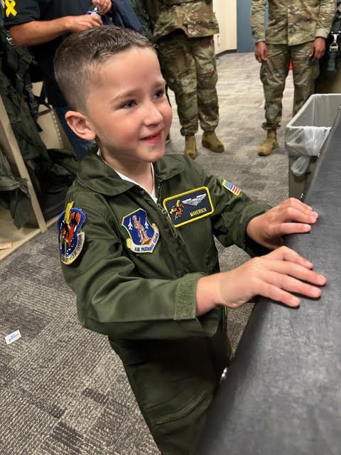 A smiling child in a green flight suit stands at a desk in a room with military personnel in the background.