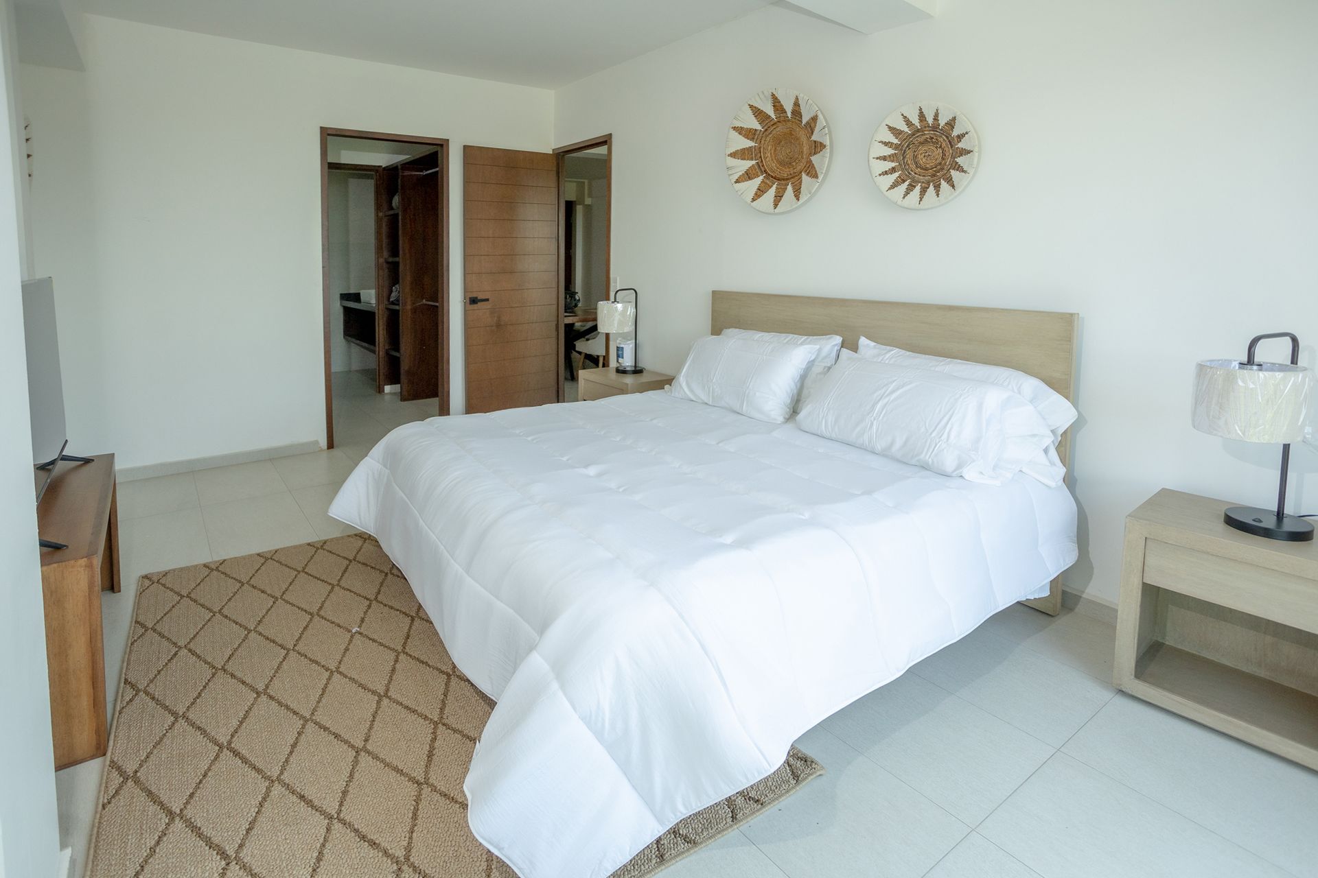 Habitación luminosa con una cama grande blanca, alfombra beige y puerta abierta que da a un pasillo.