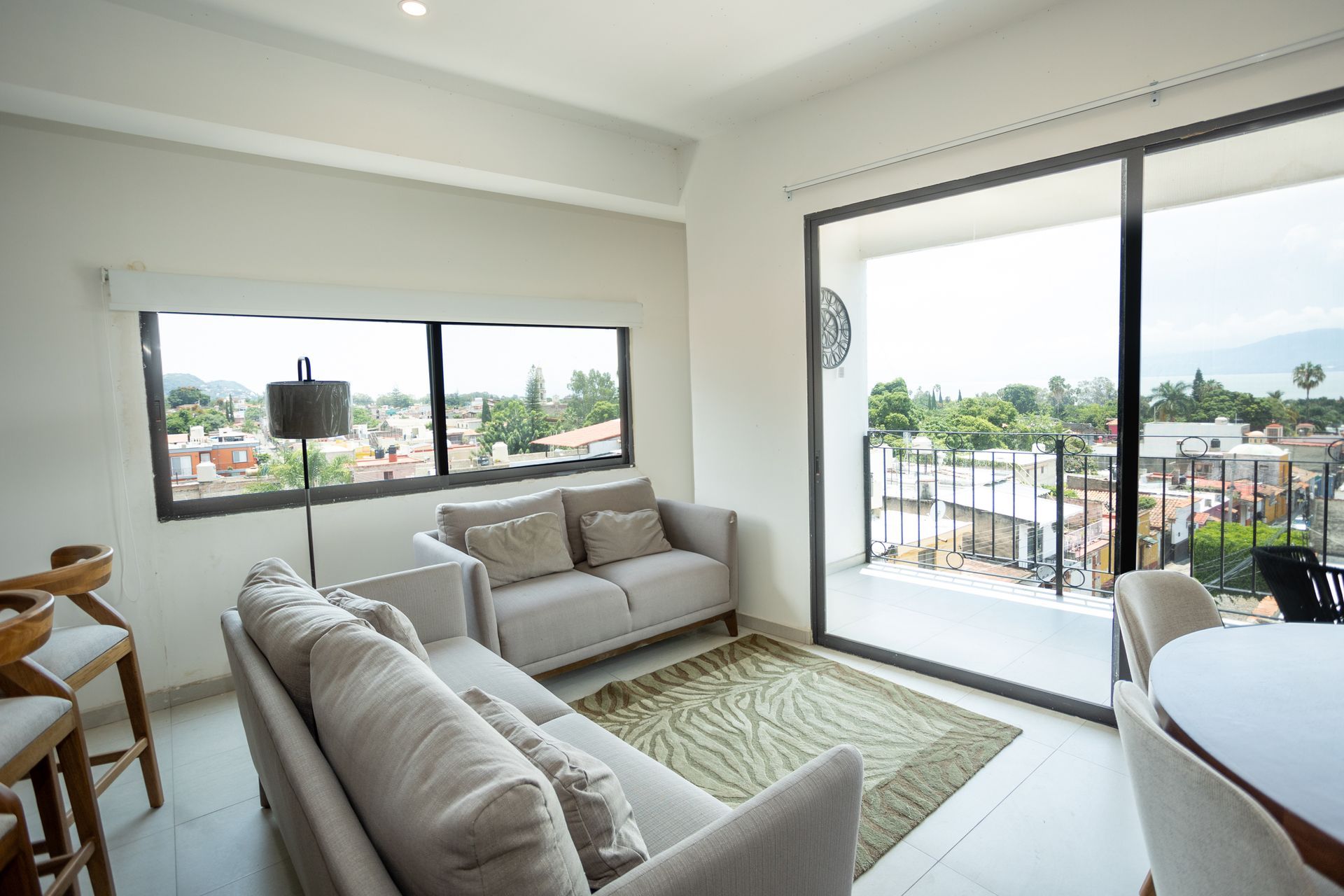 Luminoso salón con sofás beige, una alfombra y grandes ventanales con vistas a la ciudad.