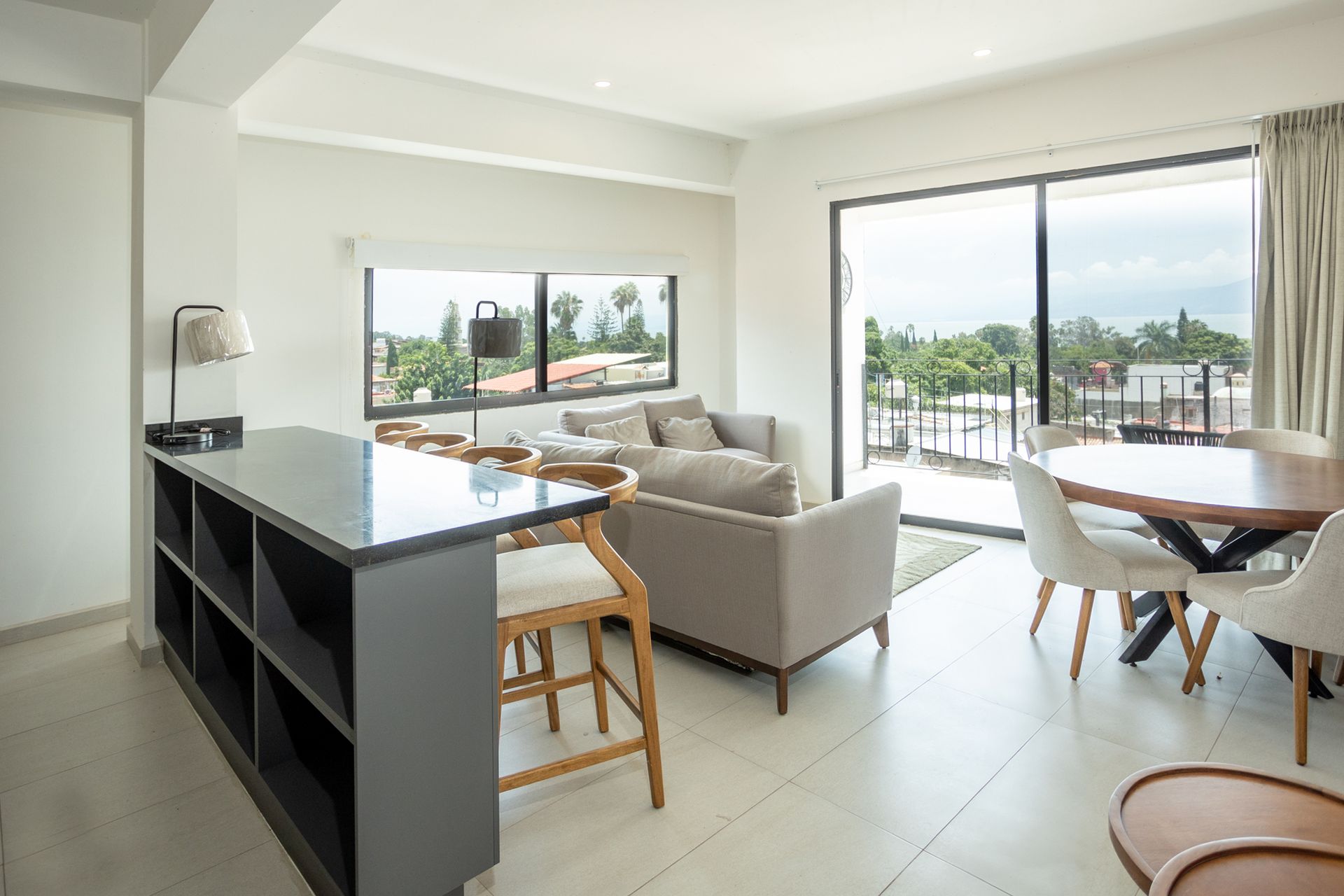 Luminoso salón-comedor diáfano con sofá beige, isla de cocina negra y puertas que dan a un balcón con vistas a los árboles.