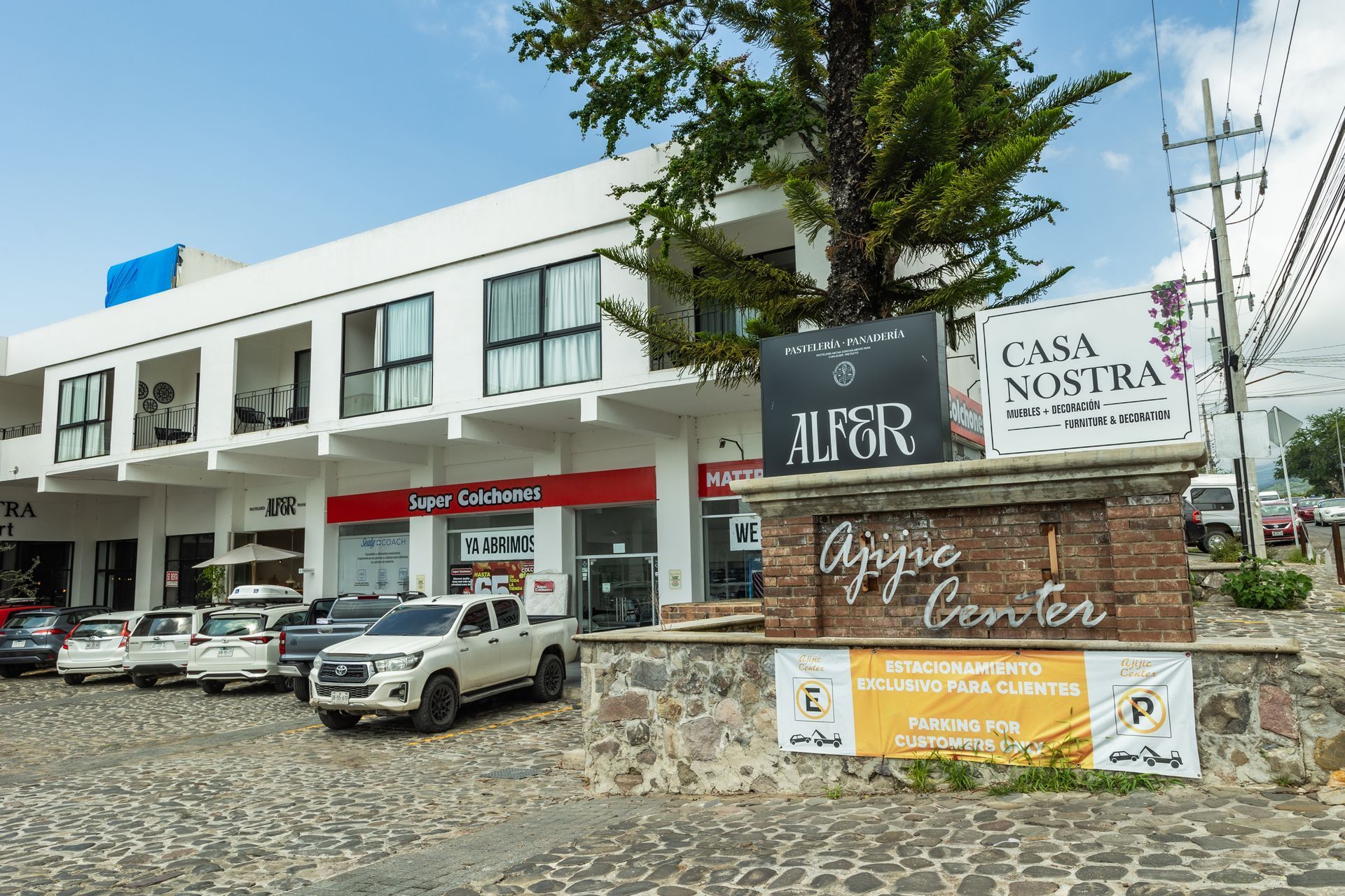Plaza comercial con coches aparcados, letrero de piedra y escaparates blancos bajo un cielo azul.