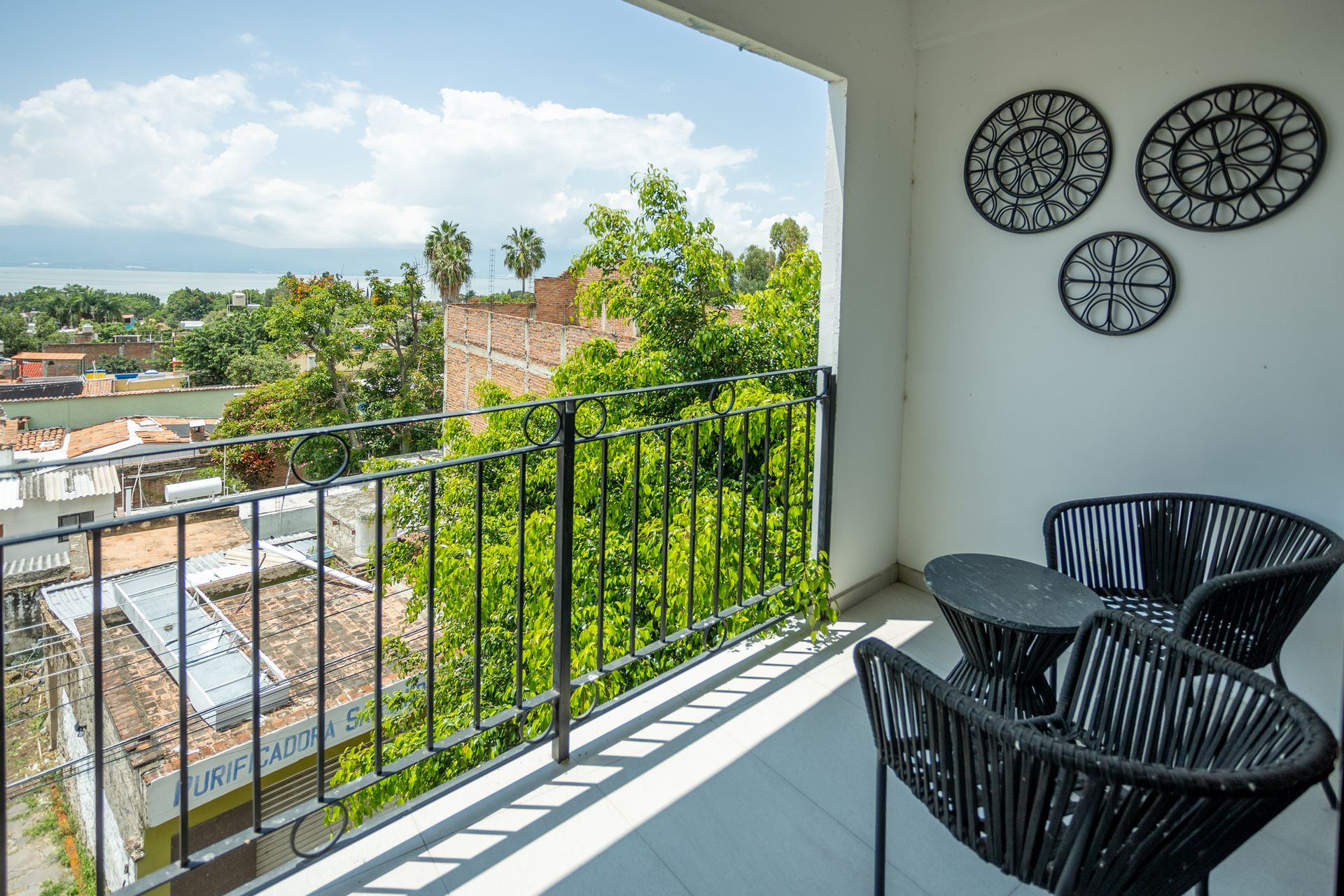 Balcón con sillas negras y decoración de pared con vistas a un barrio soleado y un paisaje verde.