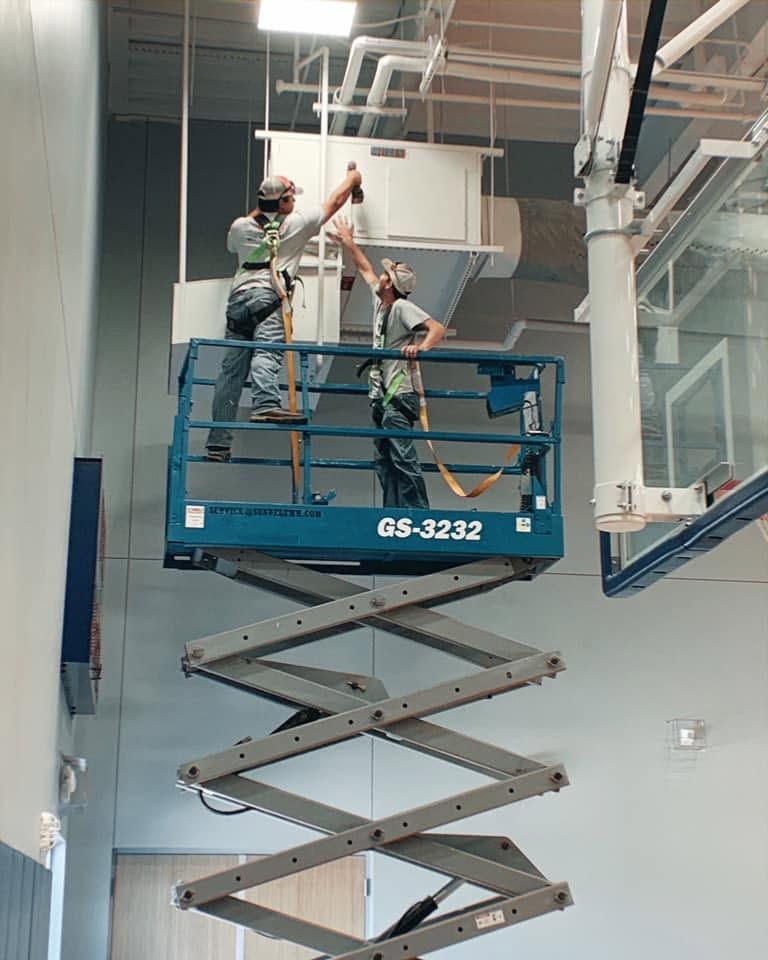 Two workers on a blue scissor lift installing equipment in an indoor industrial space