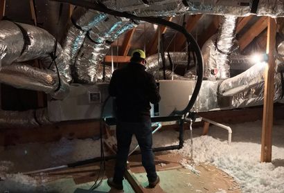 Worker in a crawl space inspecting large insulated ducts and pipes with a circular frame in the foreground.