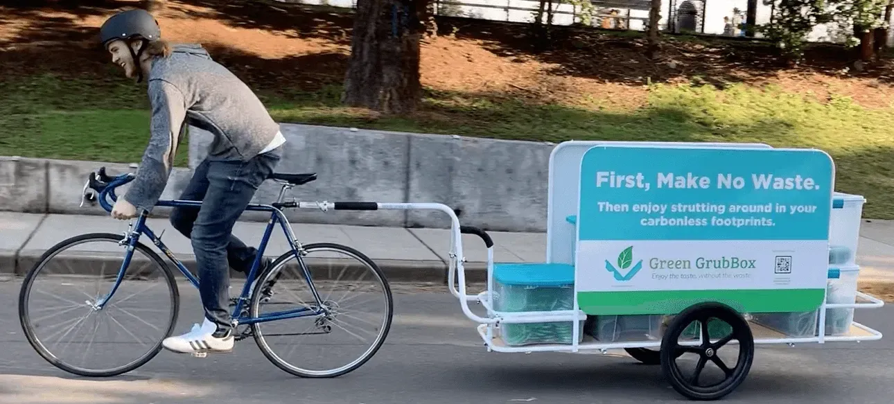 Man riding a bicycle pulling a turquoise waste bin trailer on a road