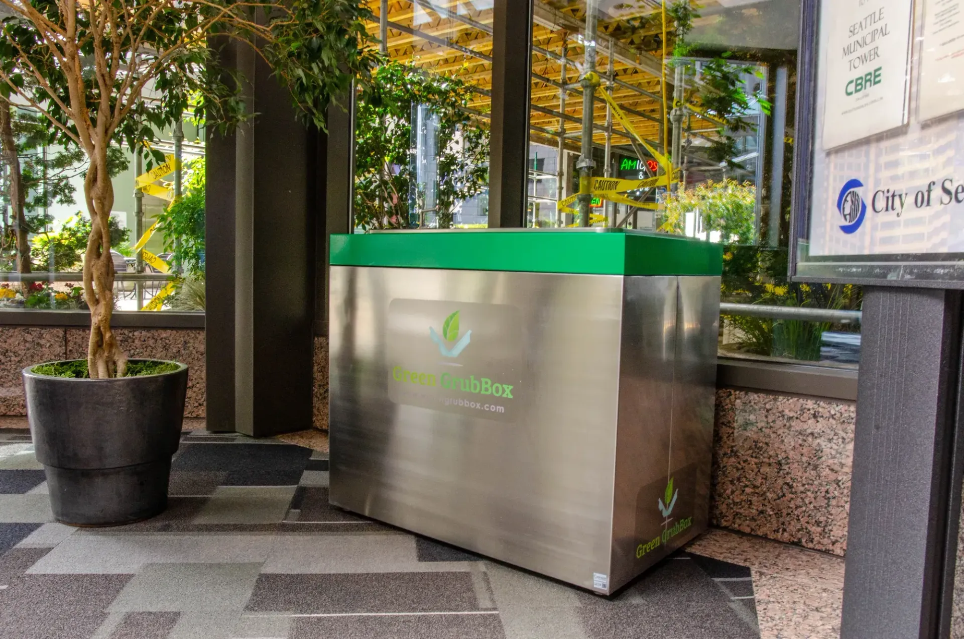 Outdoor stainless-steel recycling bin with a green lid beside a city sidewalk and trees