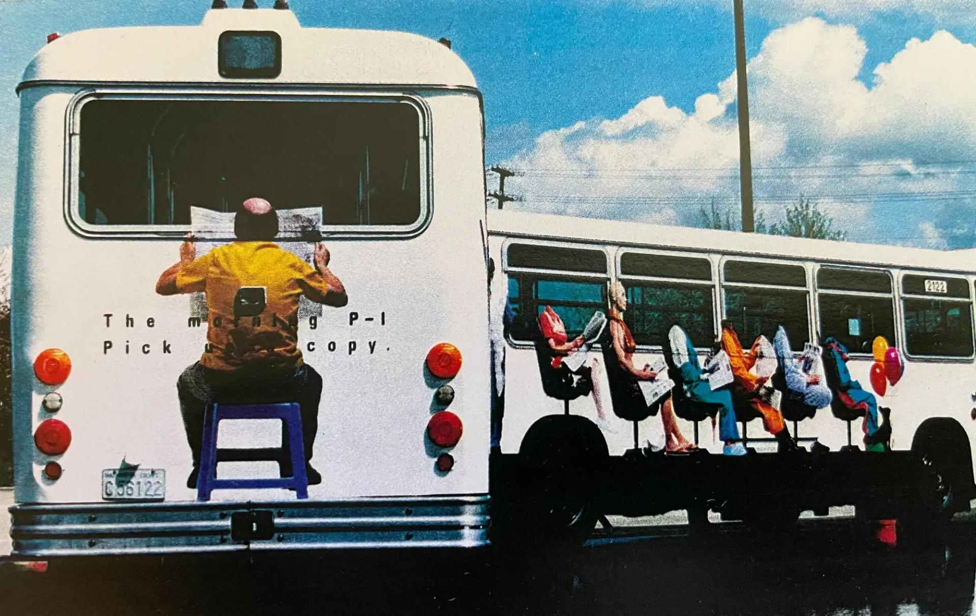 Person hanging from back of white bus beside a row of colorful seats with riders under a blue sky