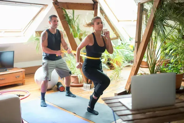 Two people doing a home workout on yoga mats in a bright loft with plants and a laptop.