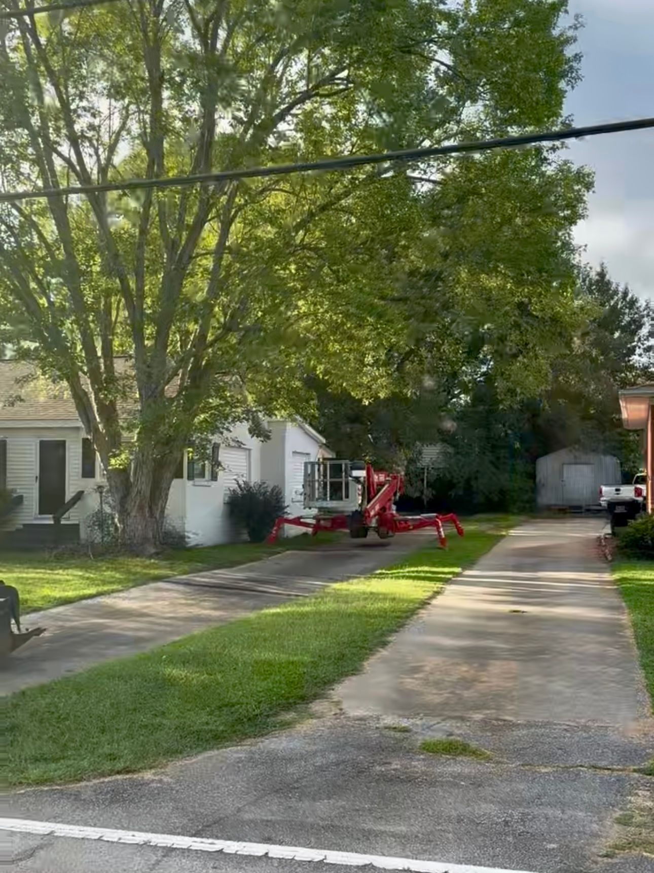 Residential driveway beside a lawn, large trees, and a red utility lift near a white house.