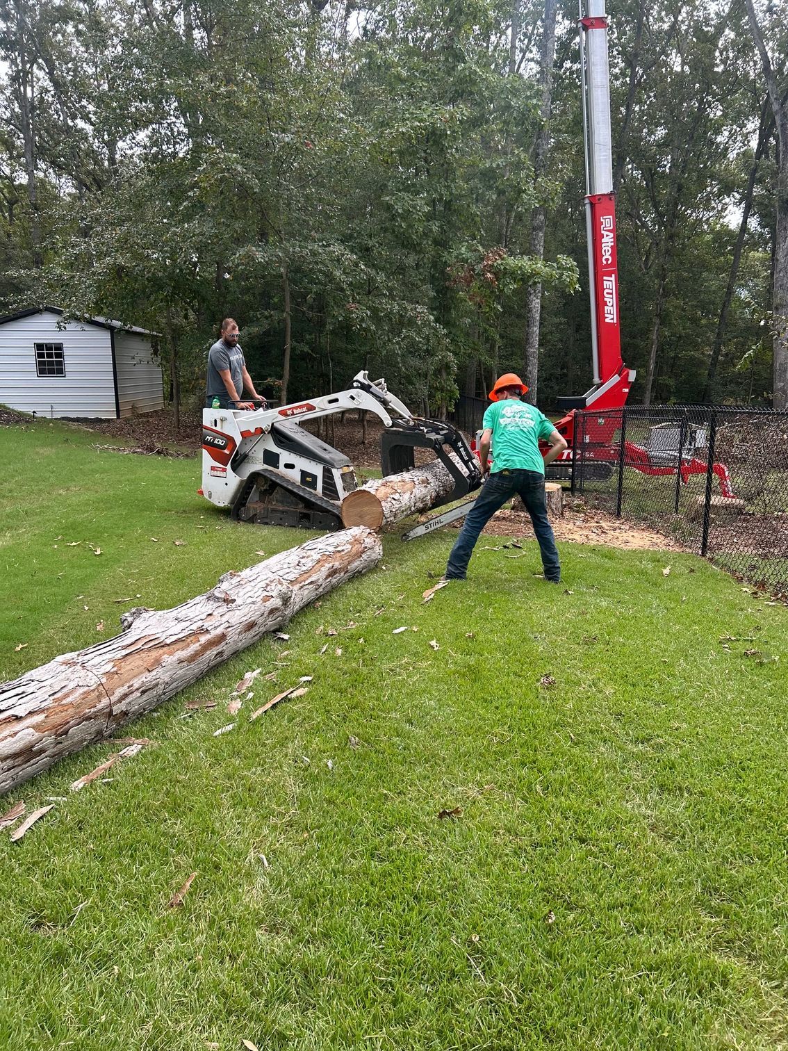 Workers with a skid steer and crane lifting a fallen tree near a house in a grassy yard