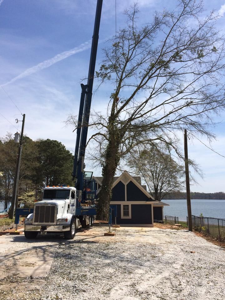 Crane truck beside a small lakeside house under construction, with a bare tree and water in the background.