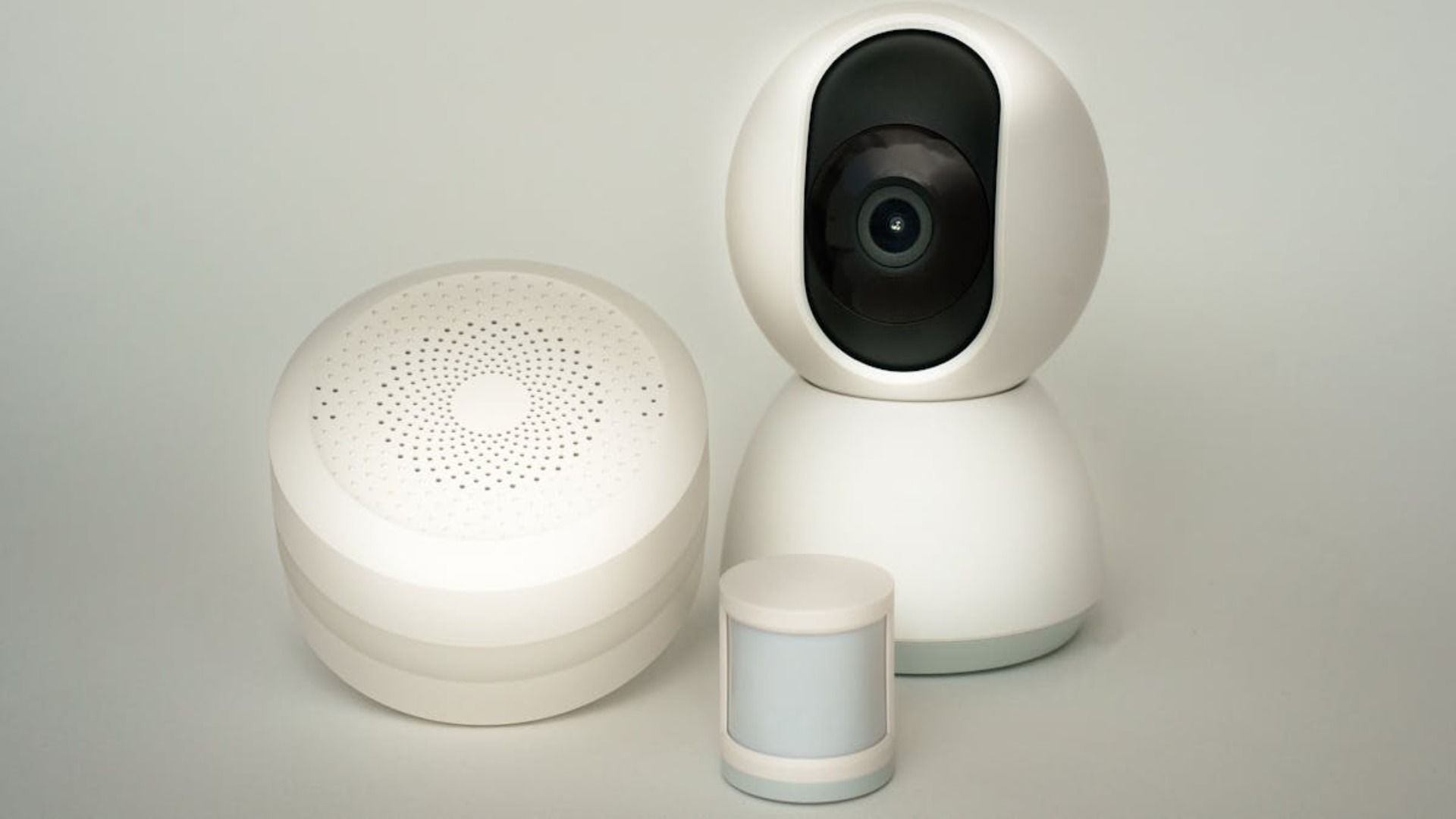 Tres dispositivos domésticos inteligentes blancos, incluyendo una cámara de seguridad, sobre un fondo claro.