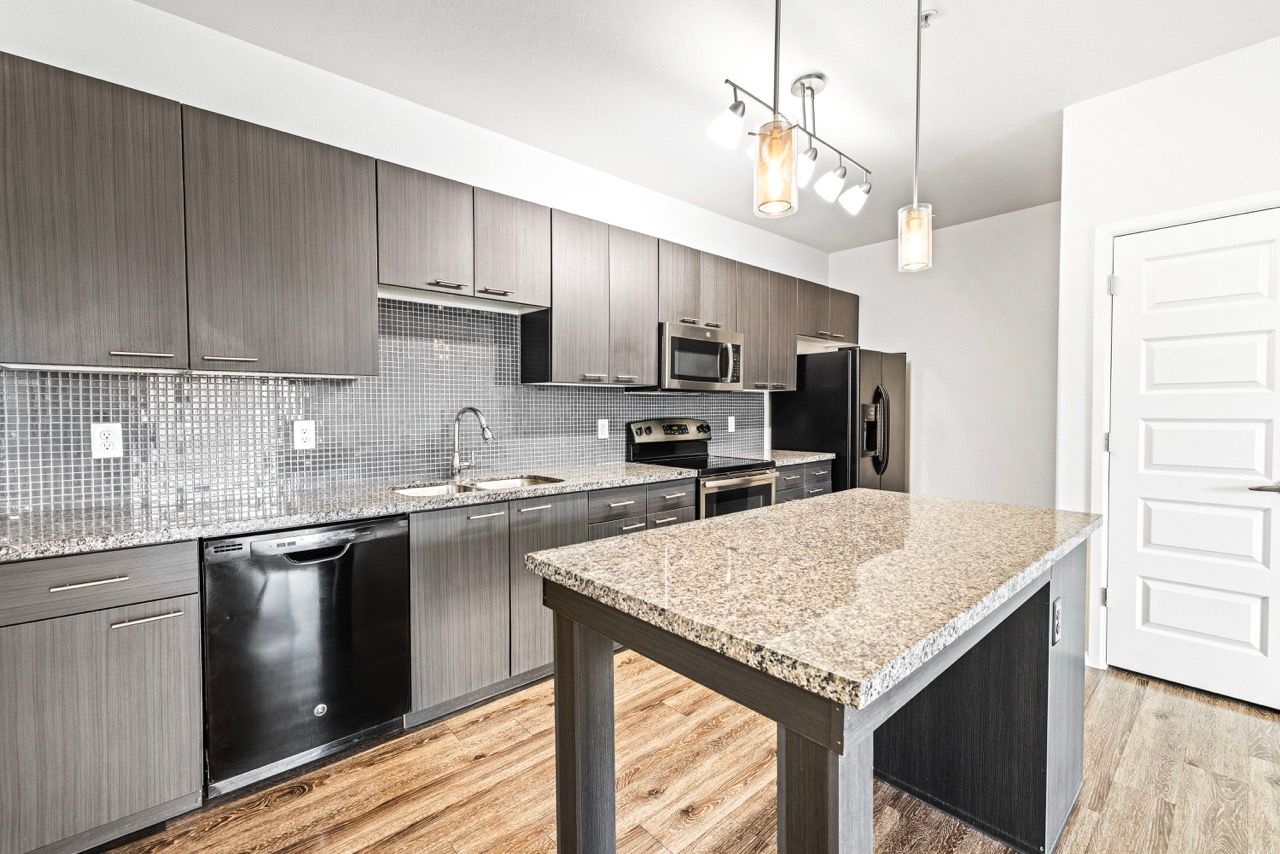 Modern kitchen with granite island, stainless steel appliances, gray wood cabinets, and a tile backsplash.