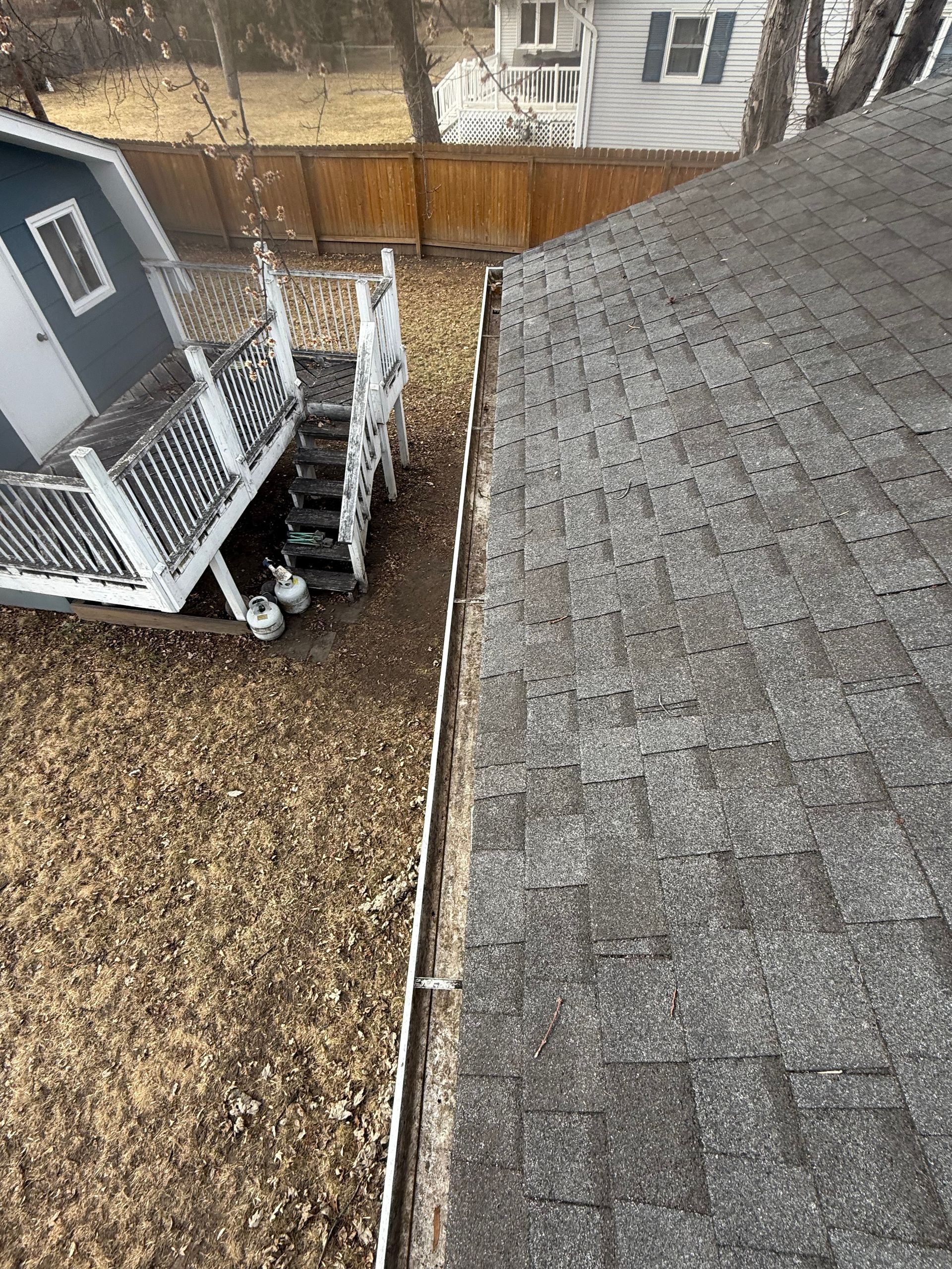 Overhead view of a house roof, yard with shed, stairs, and a wooden fence. Ground is covered in dry grass and leaves.