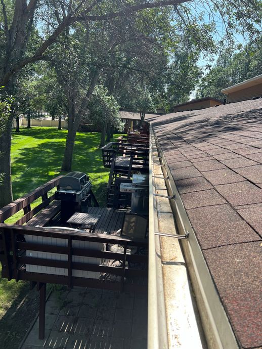 A gutter on a brown shingled roof with a wooden deck beside it, outdoors on a sunny day.