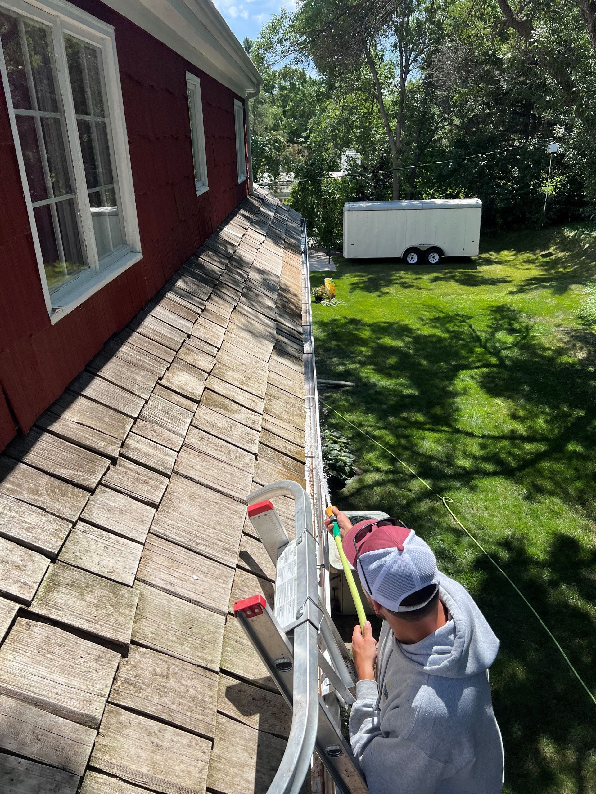 Person on a ladder measuring a gutter on a red house with a green yard and white trailer in the background.