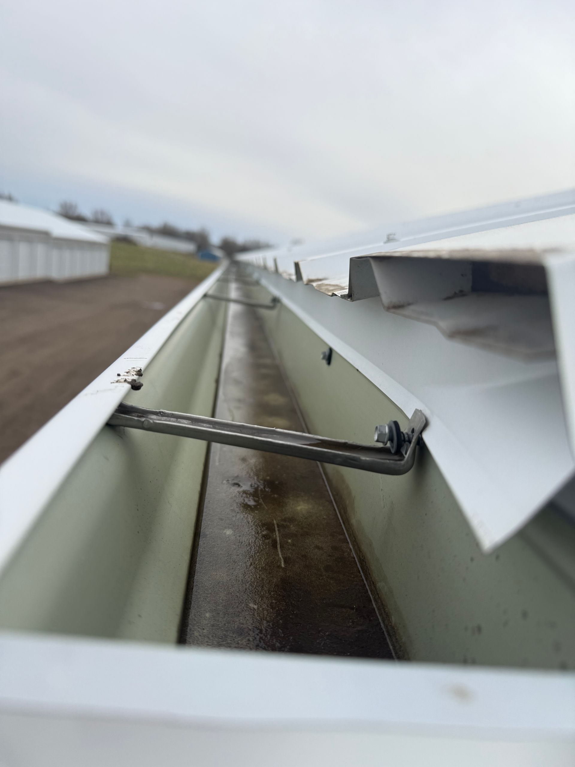 Gutter along a building roof, looking down the trough. Gray metal bar spans the gutter; cloudy sky in the background.