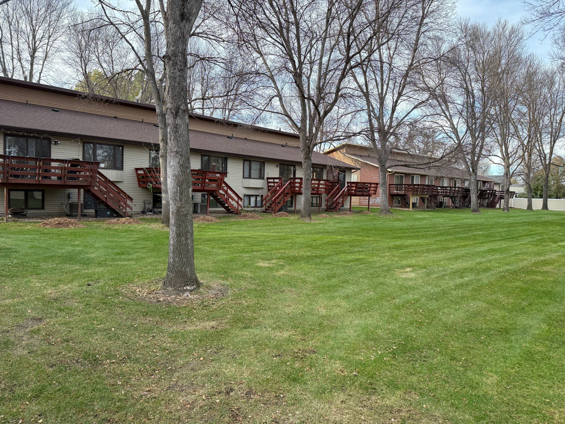 Two-story apartment buildings with brown roofs and decks, set on a green lawn with bare trees.