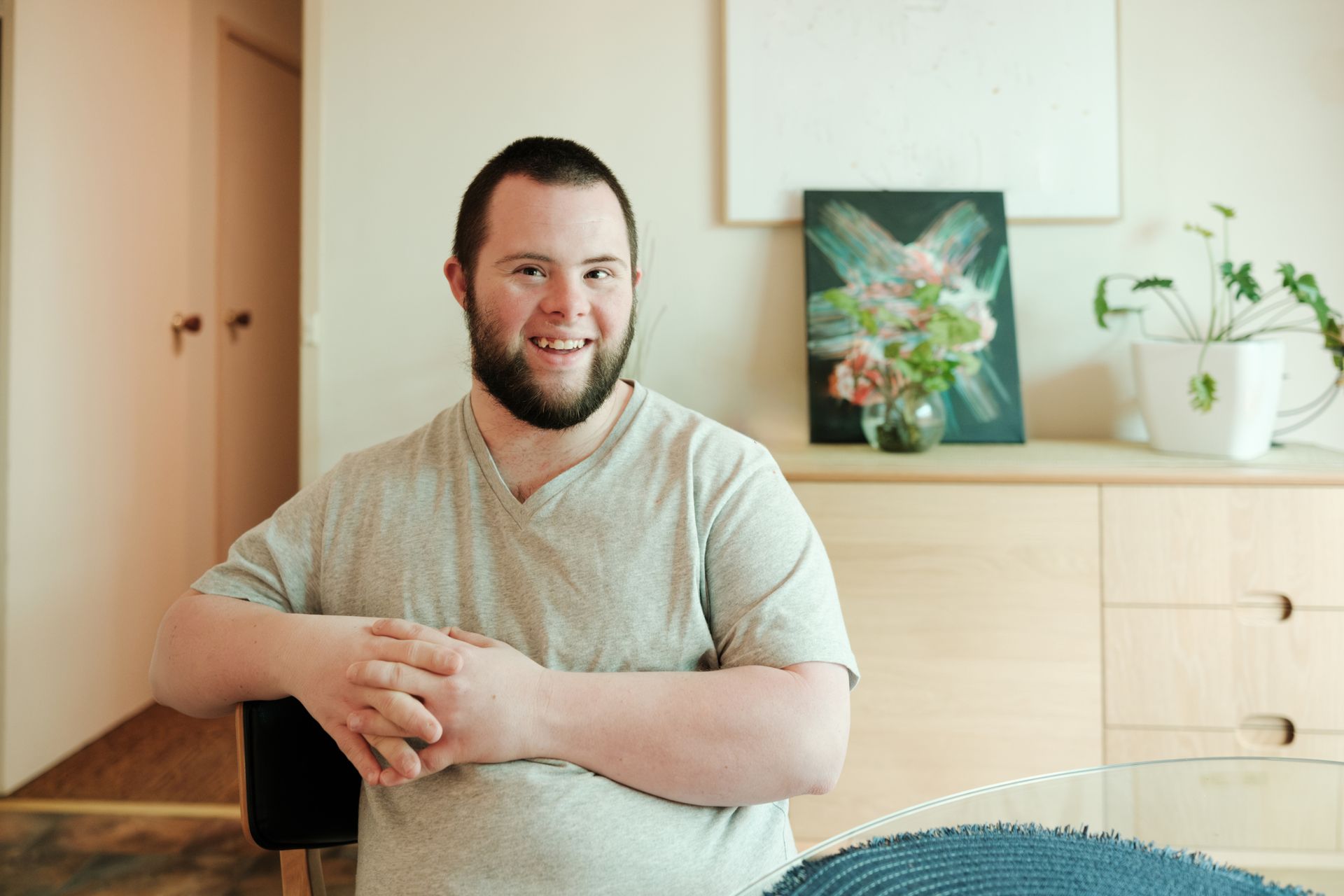 Man with Down syndrome smiling, sitting in a room, arms crossed, light-colored shirt, with a neutral expression.