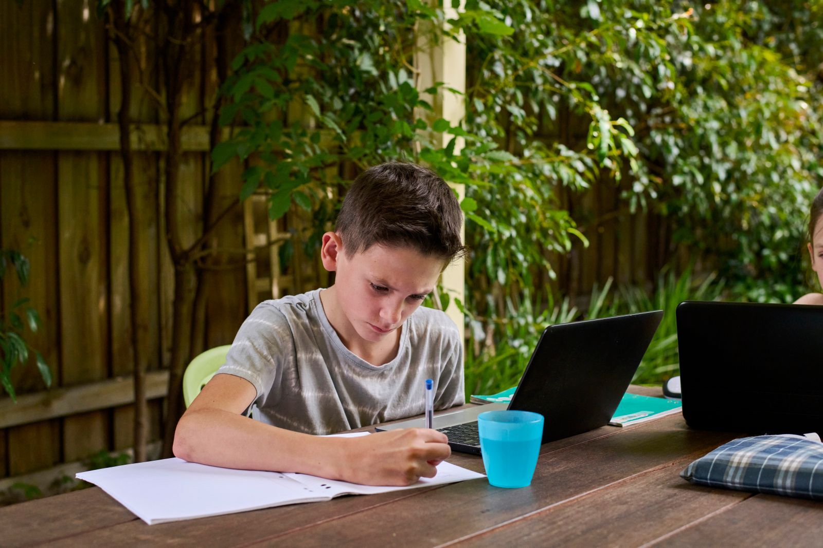 Boy writing at a table outdoors with a laptop and a blue cup.