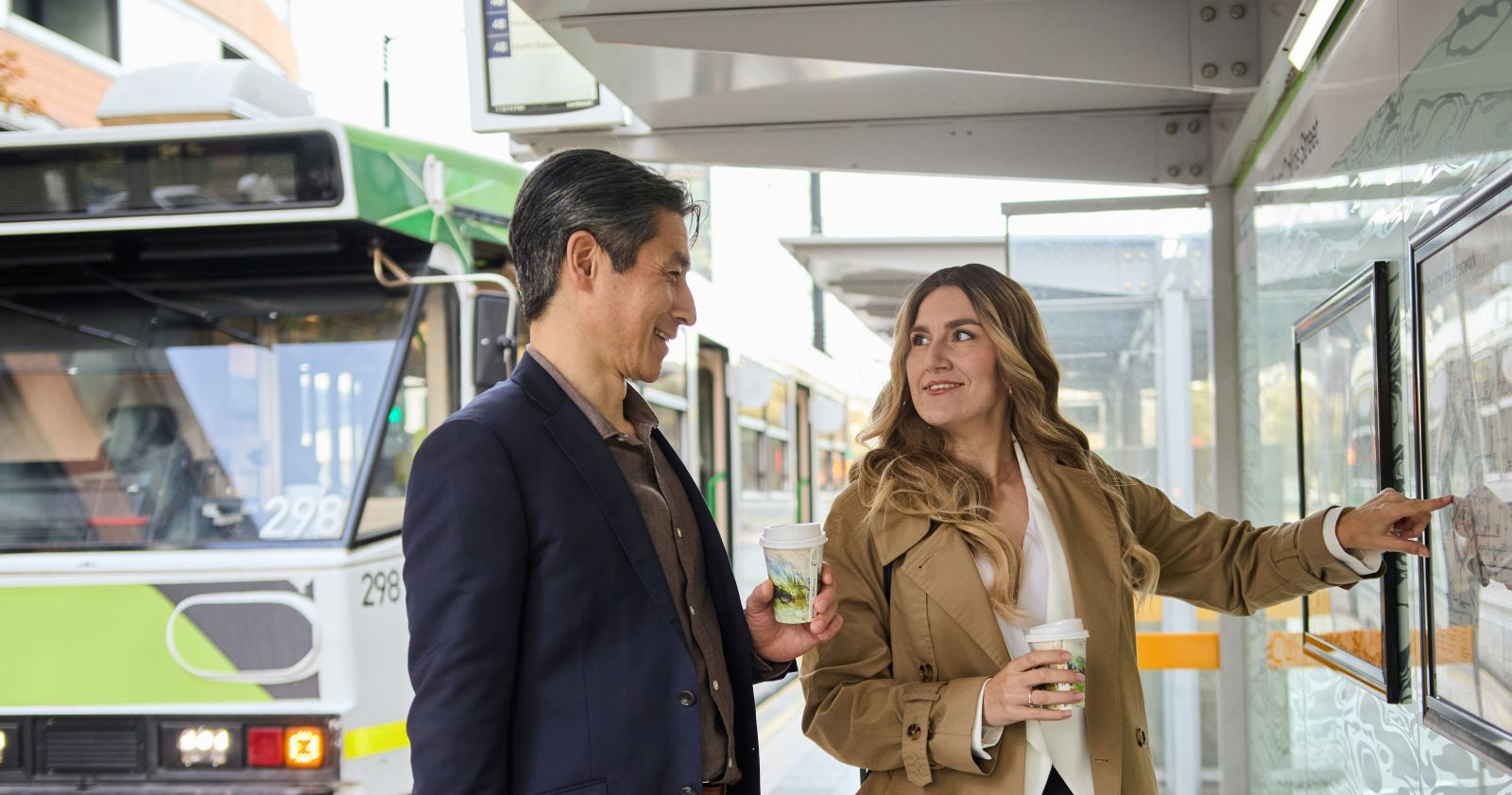 Two people stand at a bus stop, one pointing toward an arriving green and white bus.