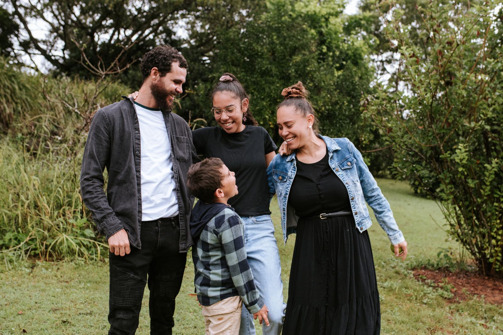 Family of five smiling outdoors: Man in cardigan, two women, and a child looking at each other, grassy area.