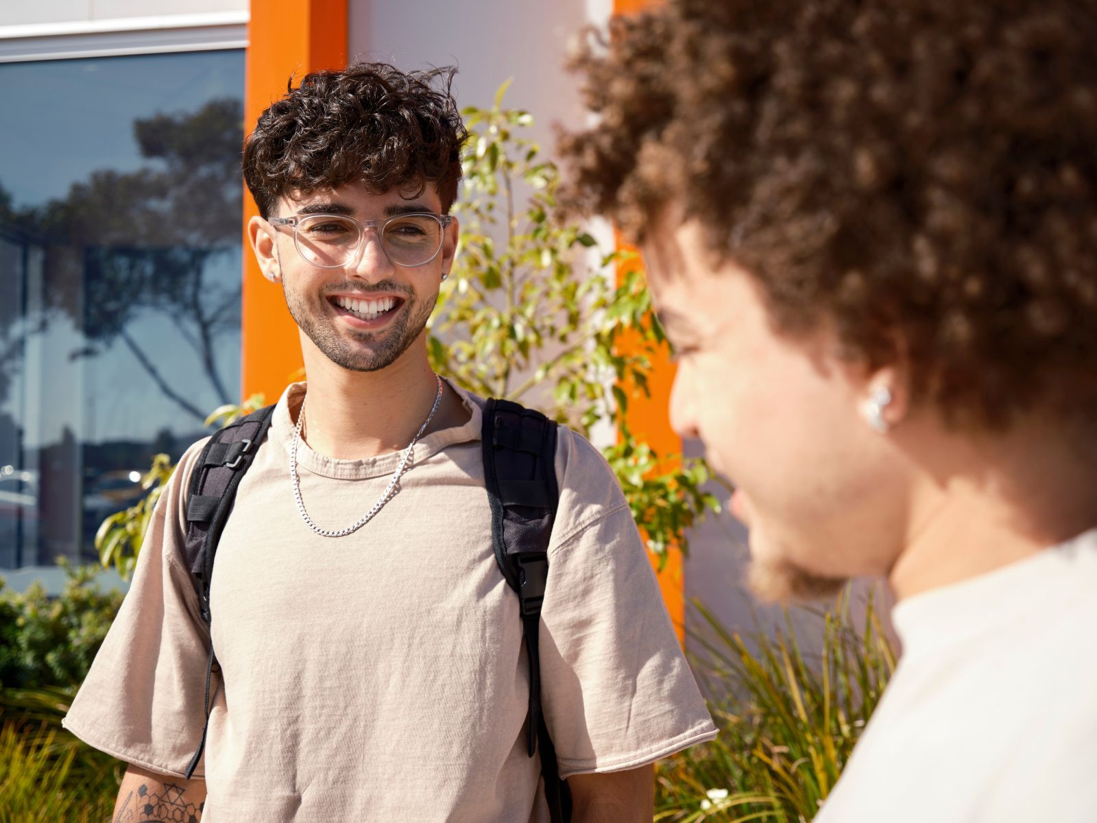 Two young men smiling at each other outdoors, one with a backpack near a building with orange accents.