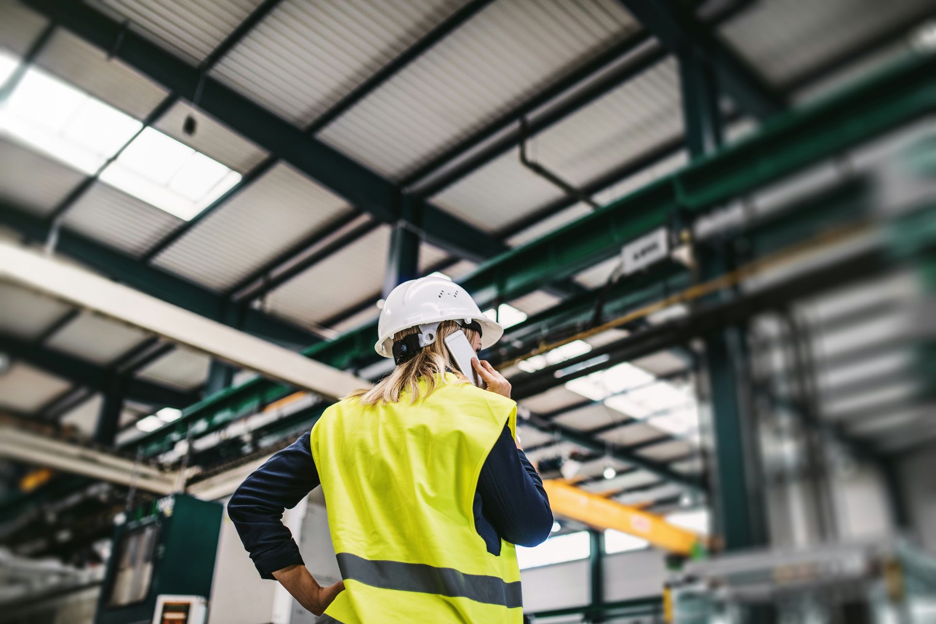 A person in a hard hat and high-visibility yellow vest stands in a factory, talking on a mobile phone.