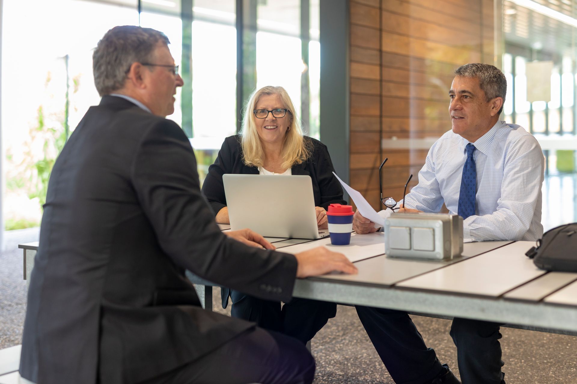 Three coworkers in a bright meeting room talking around a table with a laptop and documents