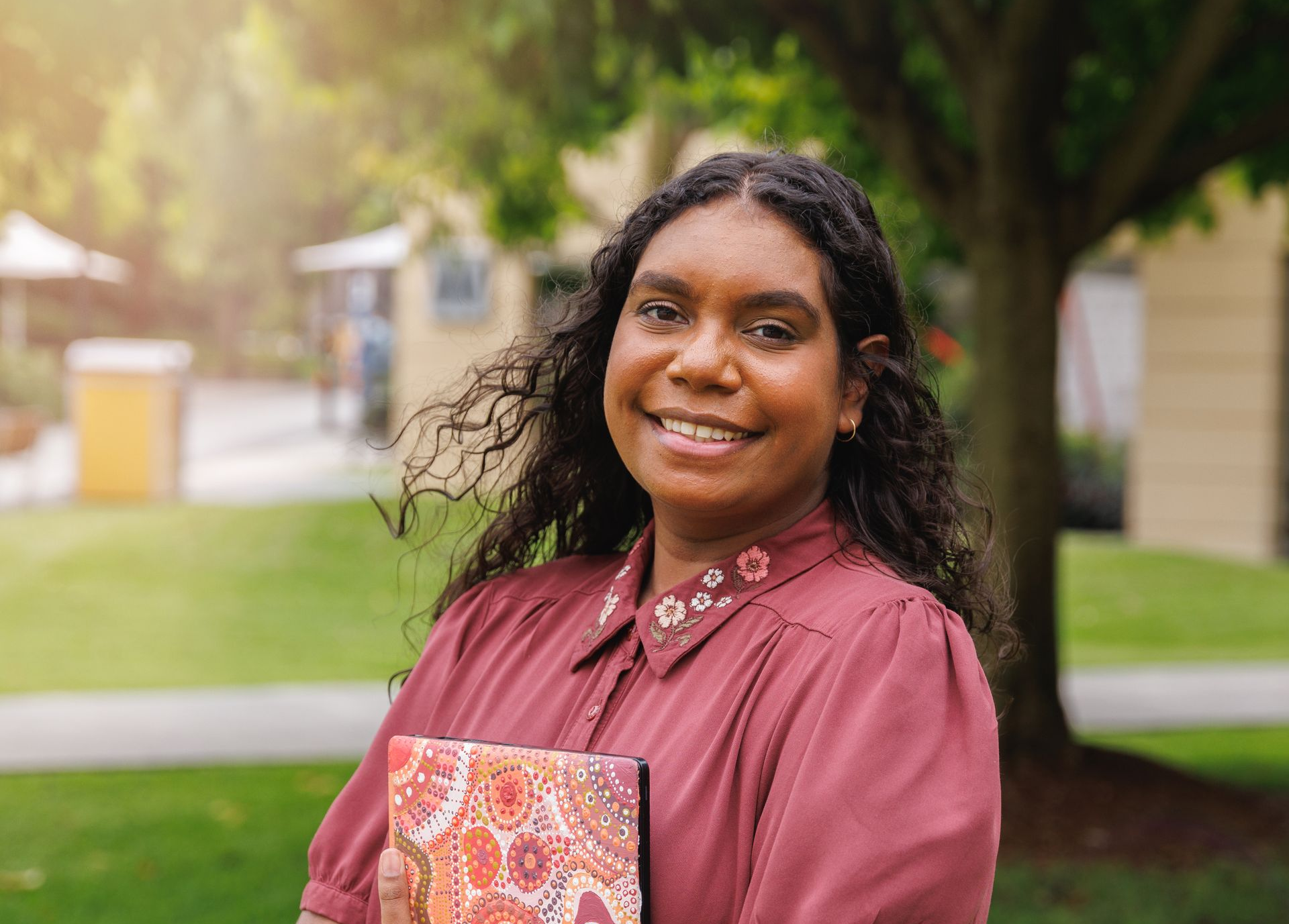 Woman in maroon shirt and pink pants smiles, holding a patterned book, standing in a park.