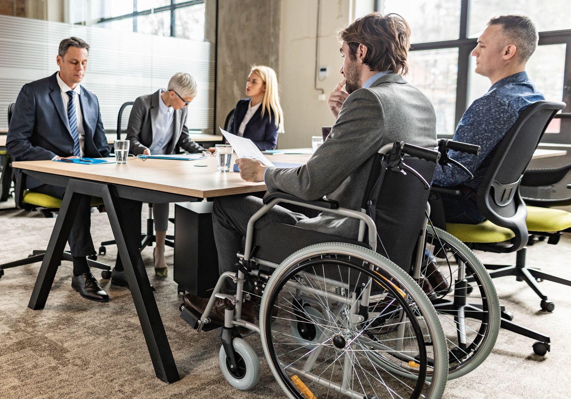 Meeting room discussion with people seated around a table, including one person in a wheelchair.
