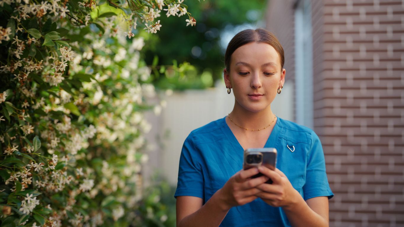 Person in a blue shirt looking at a phone outdoors beside flowering bushes and a brick wall