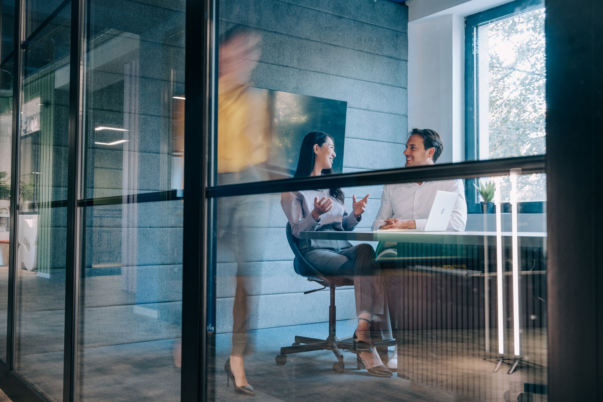 People meeting in a glass-walled office, with a blurred person walking past in the foreground.