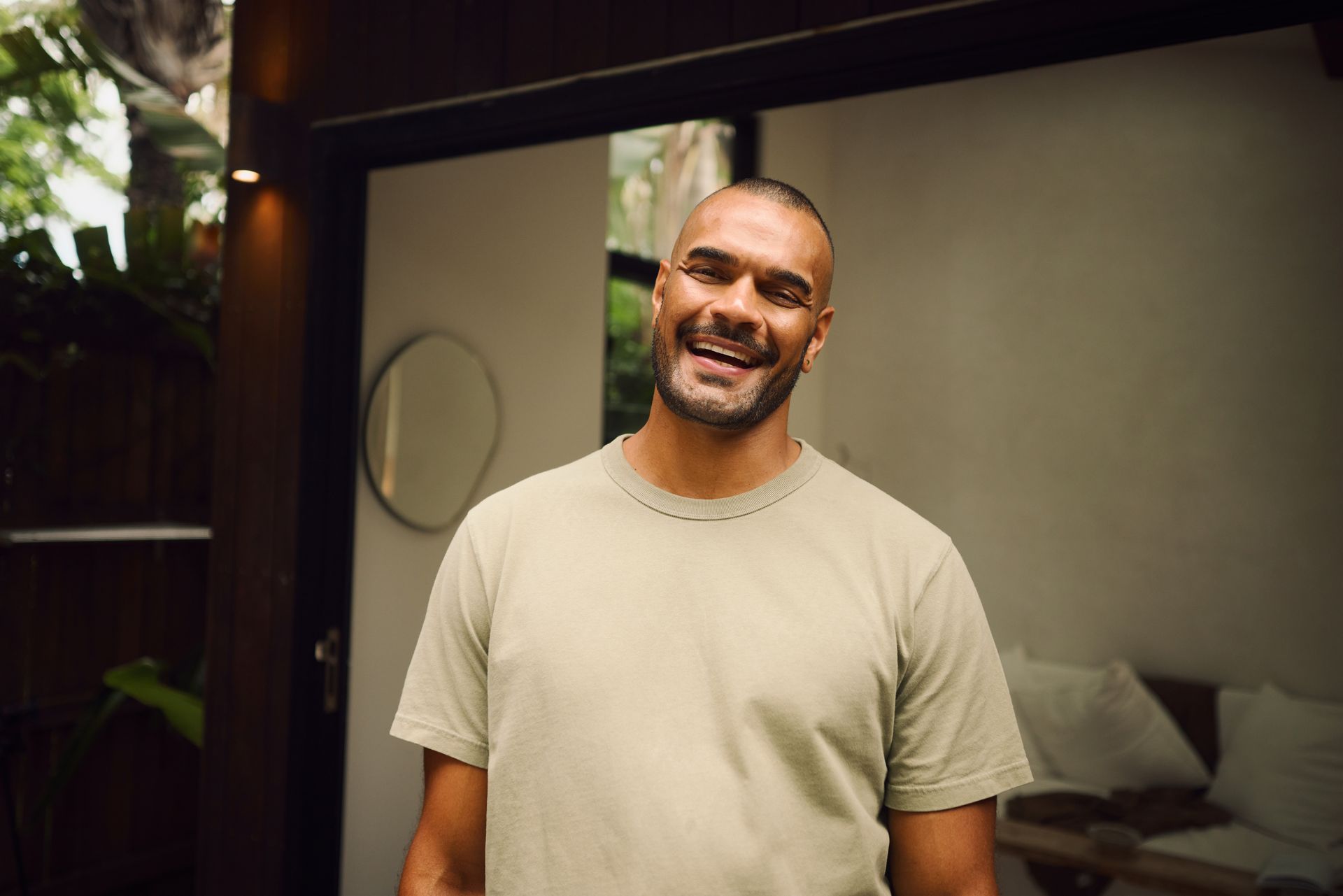 Man with shaved head smiling, wearing a light green t-shirt, standing outdoors in front of a white door.