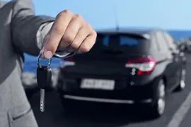 Hand holding car key fob in front of a parked black car on a road