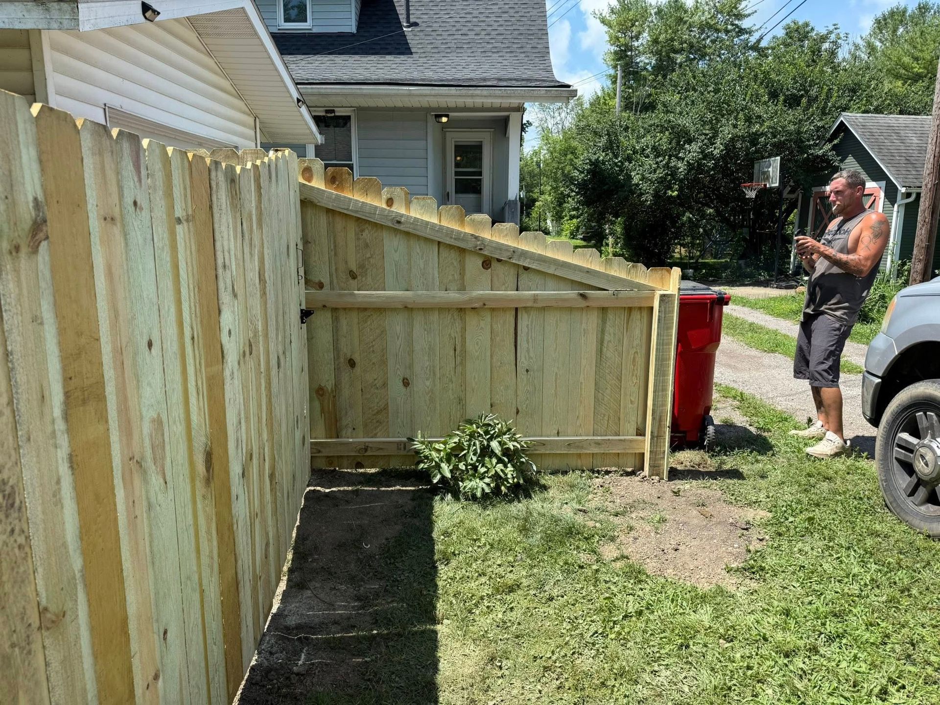 Man standing next to a wooden fence and red trash bin in a backyard on a sunny day.