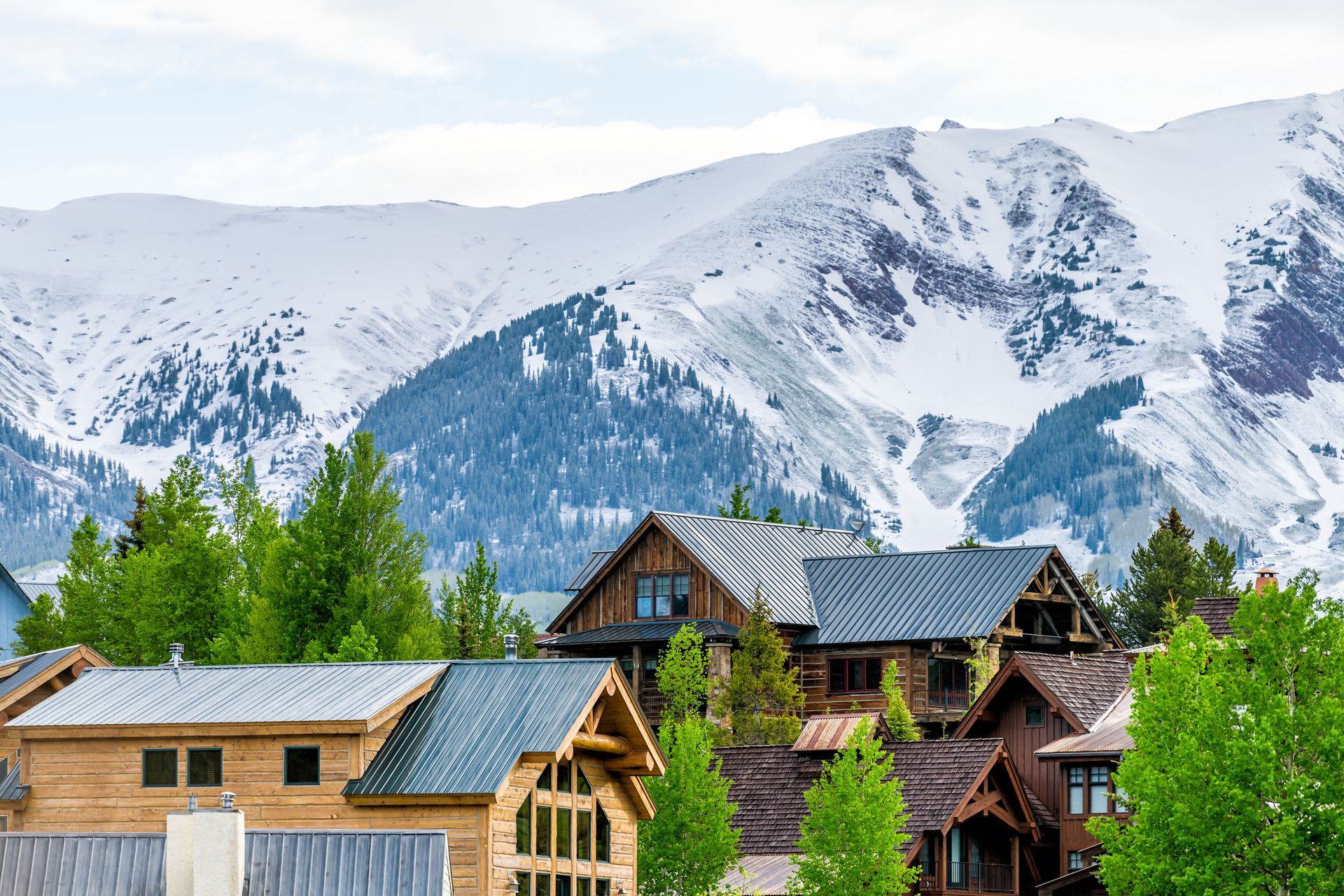 Wooden houses nestled below snow-covered mountains and lush green trees.