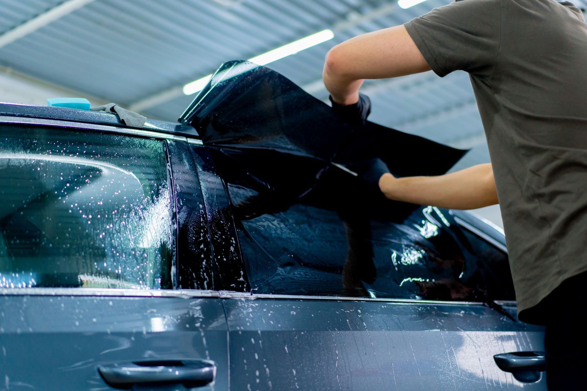 A person wearing black gloves carefully applies dark window tinting film to a wet car window inside a garage.