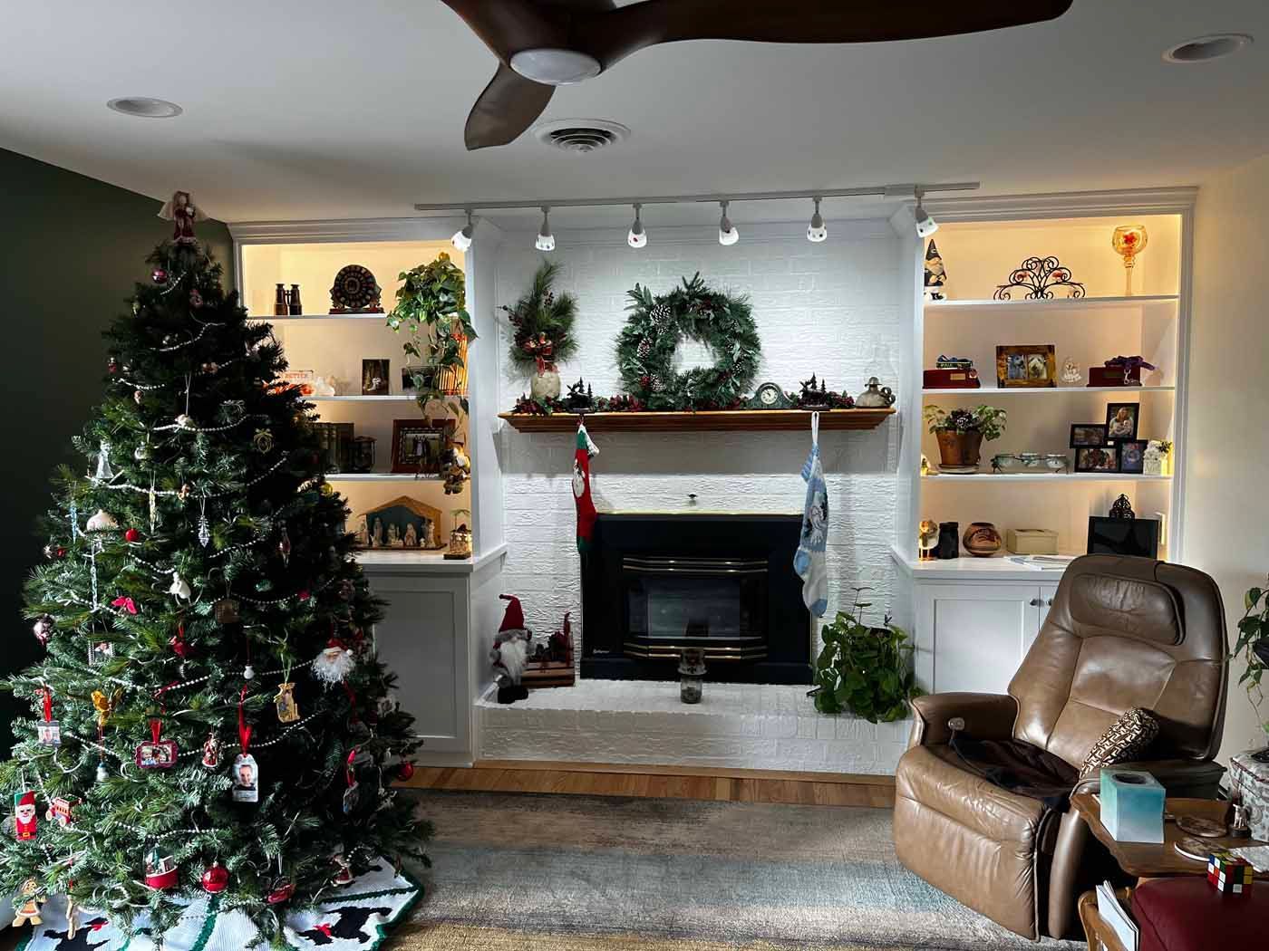 Festively decorated living room with Christmas tree, fireplace, wreath, shelves, and leather chair