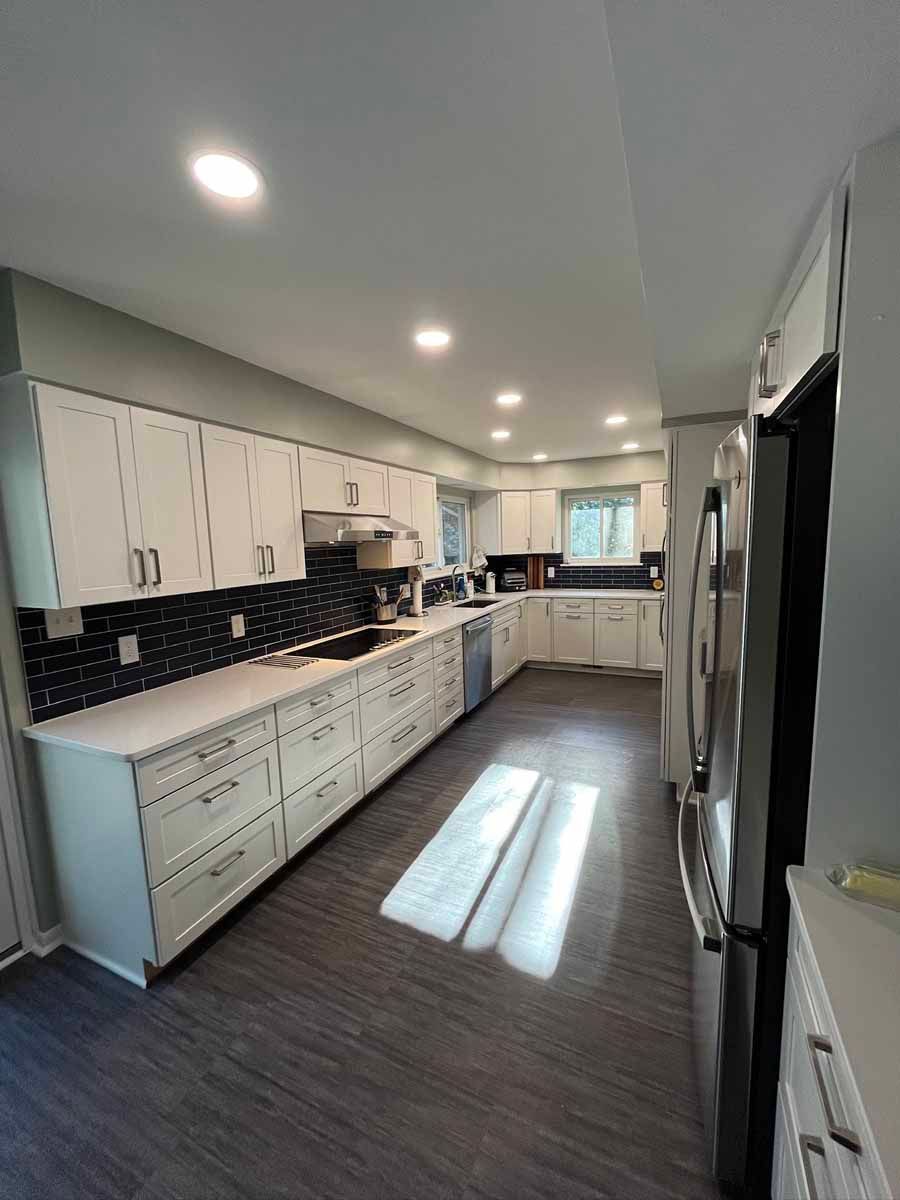 Bright white galley kitchen with black backsplash, stainless fridge, and sunlight on wood floors