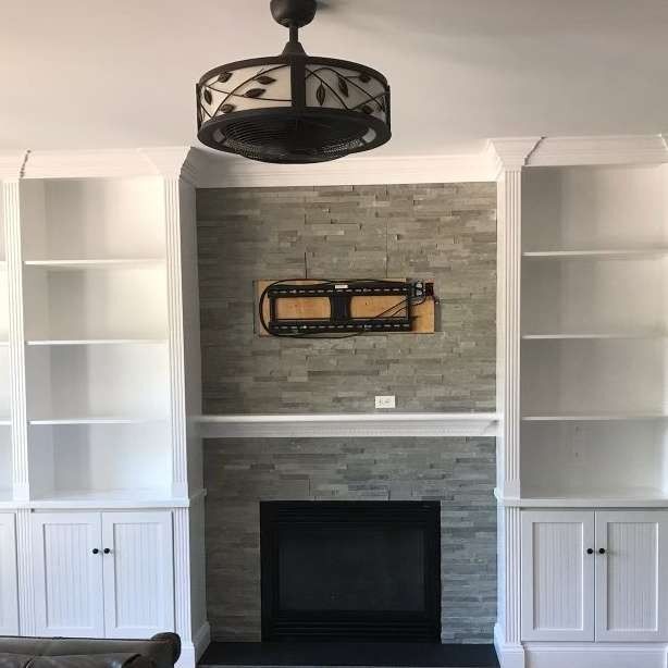 Built-in white shelves and cabinets flanking a gray stone fireplace with a black mantel and ceiling fan.