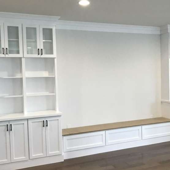 White built-in cabinets and shelves beside a long window seat with a wood top in a bright room.