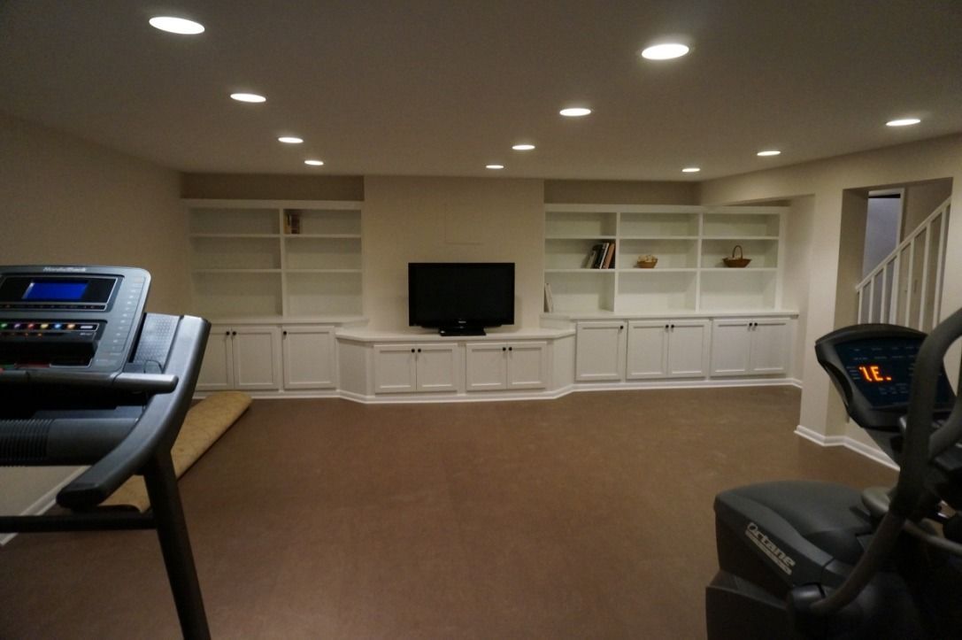 Finished basement family room with white built-in cabinets, wall TV, brown carpet, and workout equipment on both sides