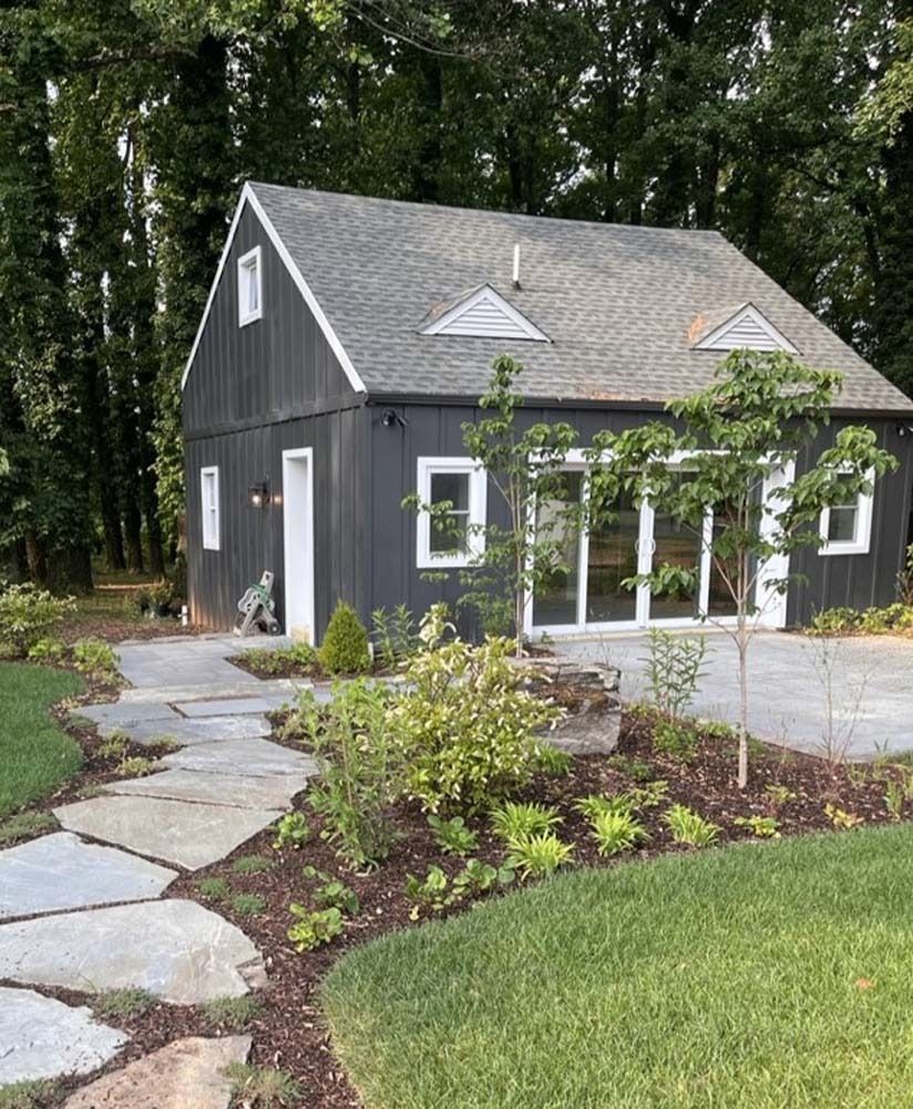 Gray cottage with white trim, stone path, and landscaped yard beside tall trees