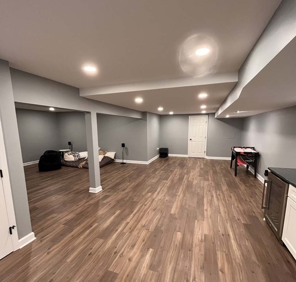 Spacious empty basement room with gray walls, wood flooring, recessed lights, and a bed in the corner