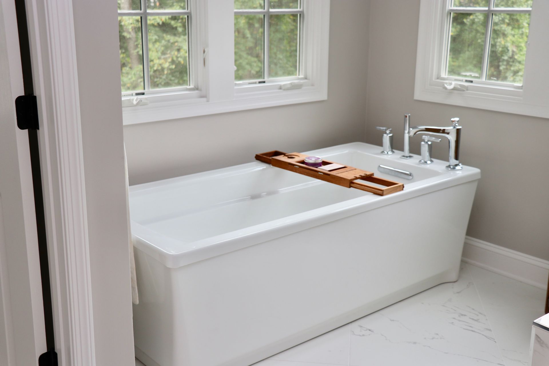 White bathroom with a soaking tub, wooden bath tray, and sink beneath three windows.