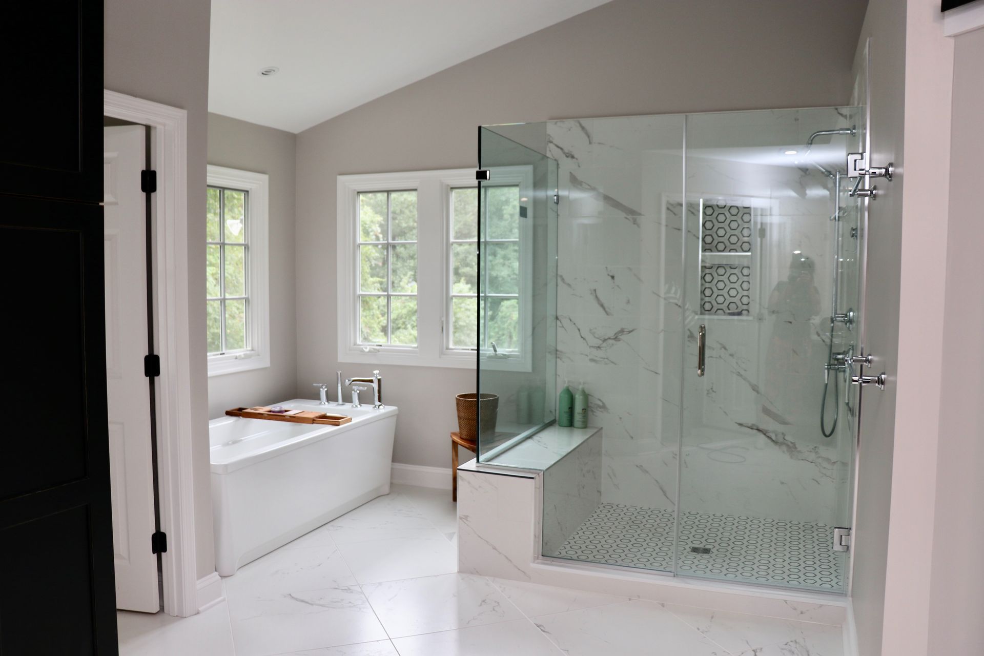 Bright white bathroom with a soaking tub, glass shower, and windows overlooking greenery.