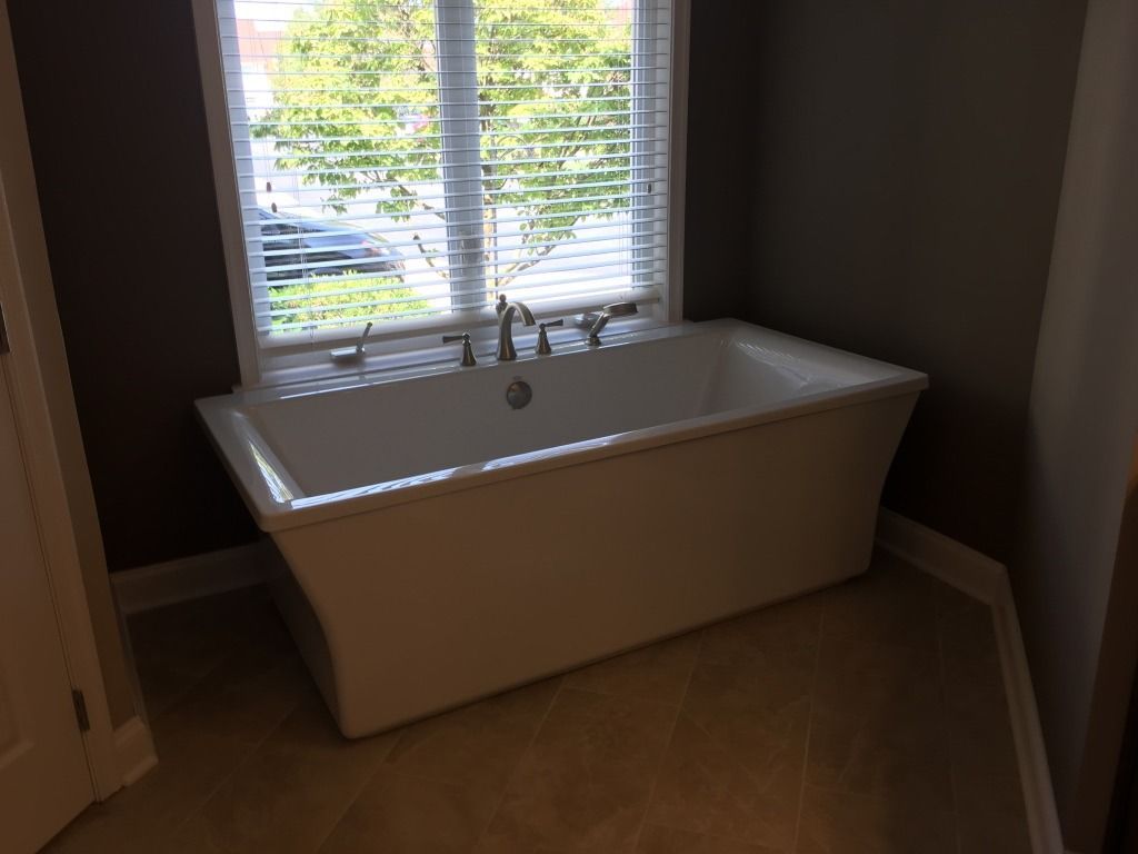 Bathroom with a freestanding white bathtub beneath a window with blinds and a sink faucet.